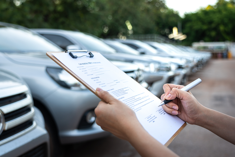 Car rental concept with fleet of cars lined up and person filling out rental agreement on a clipboard to visualize one of the cheapest ways to rent a car 