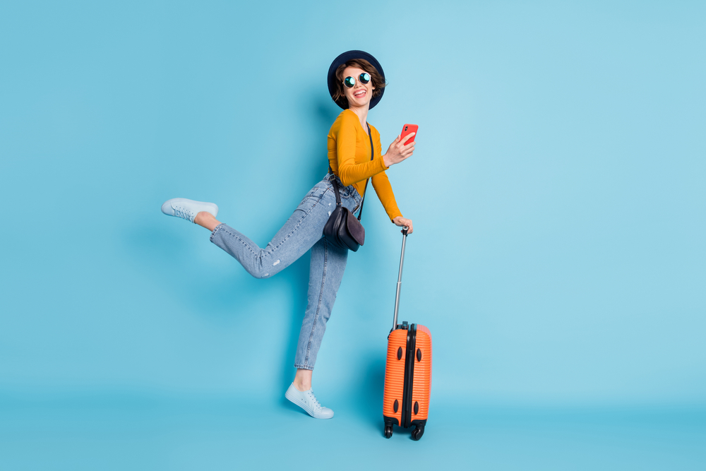 Woman in yellow shirt kicks one leg up with luggage and sunglasses on for a frequently asked questions section on how far in advance should you book a flight