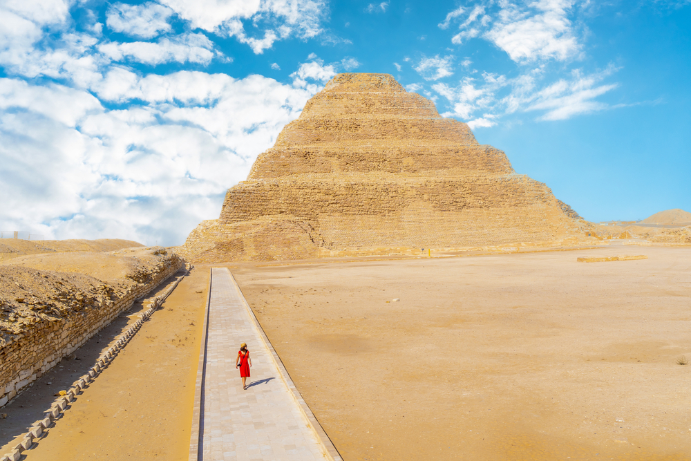 Thin young woman in a bright red dress walking on a long path through the desert that leads to the tiered pyramid in Saqqara, a top pick for the best places to visit in Egypt