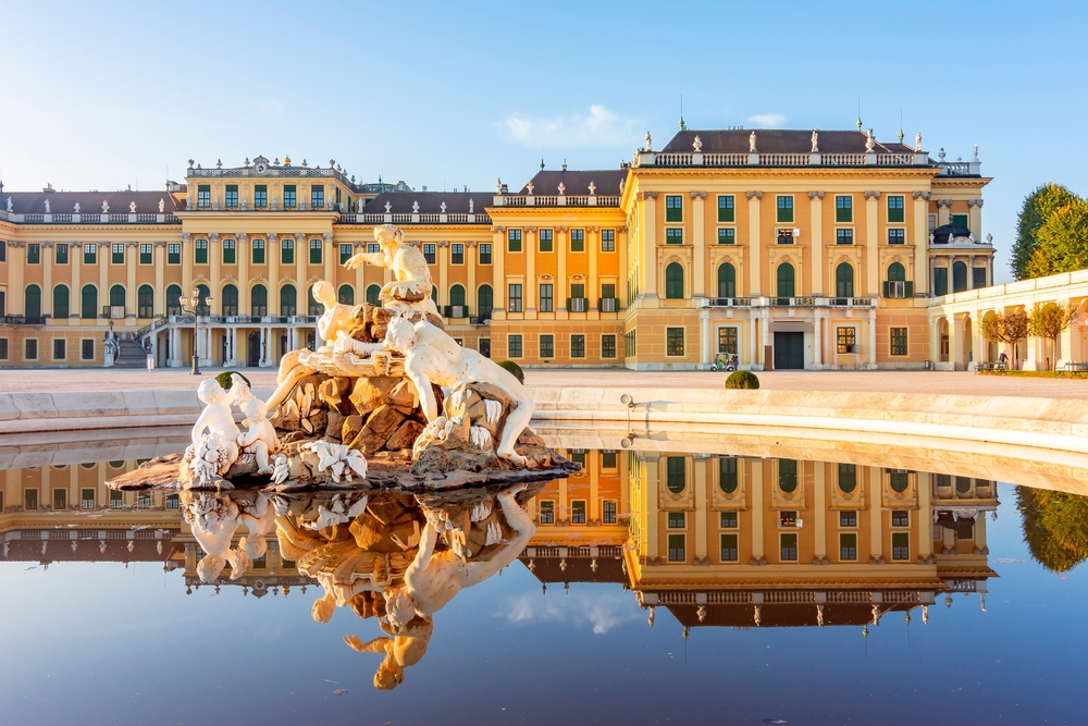 Schönbrunn Palace, one of the must-see places to visit in Austria, pictured with the yellow building reflecting all around the marble sculpture in the pool outside of it