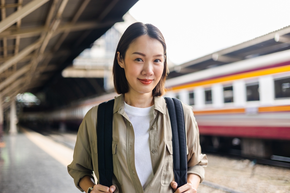 Asian young woman smiles while holding the straps of one of the best travel backpacks for women in front of a train station terminal