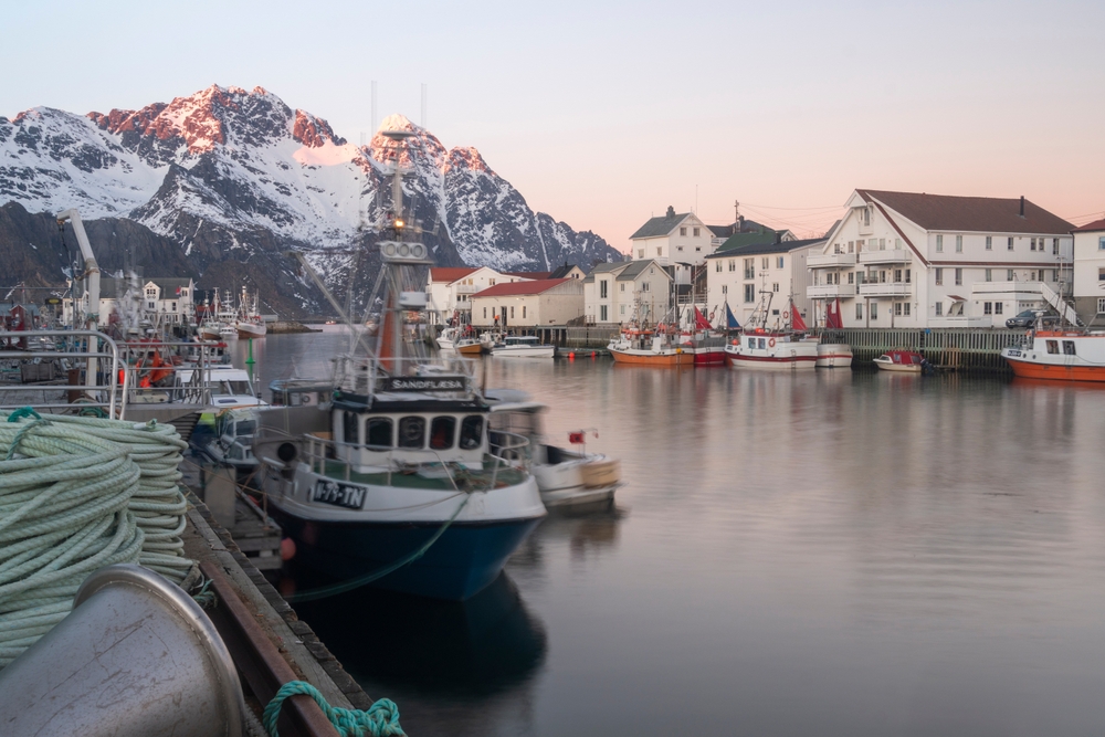 Small harbor town of Austvagoya, one of the best places to visit in Norway, pictured with a boat docked at the harbor and snow-capped mountains all around
