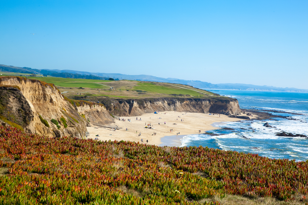 Tourists on the white sand of Half Moon Bay with waves lapping the water for a guide to the best day trips from San Francisco