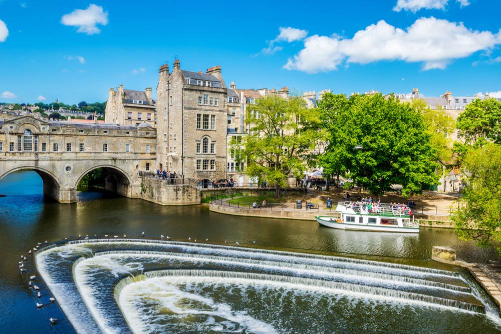 Water flowing over the decorative fountain in Bath, one of the best places to visit in England, while a tour boat drives by going under the bridge