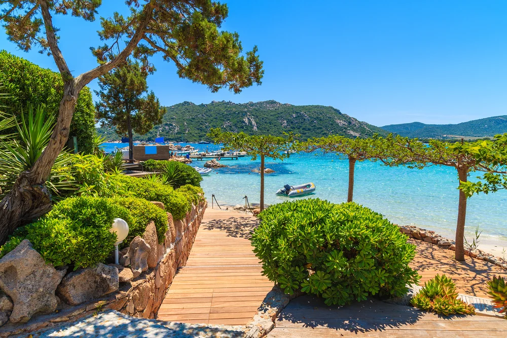 Wooden boardwalk between trees leading to the deep blue water off the Santa Biulia Beach in Corsica, one of the best places to visit in France
