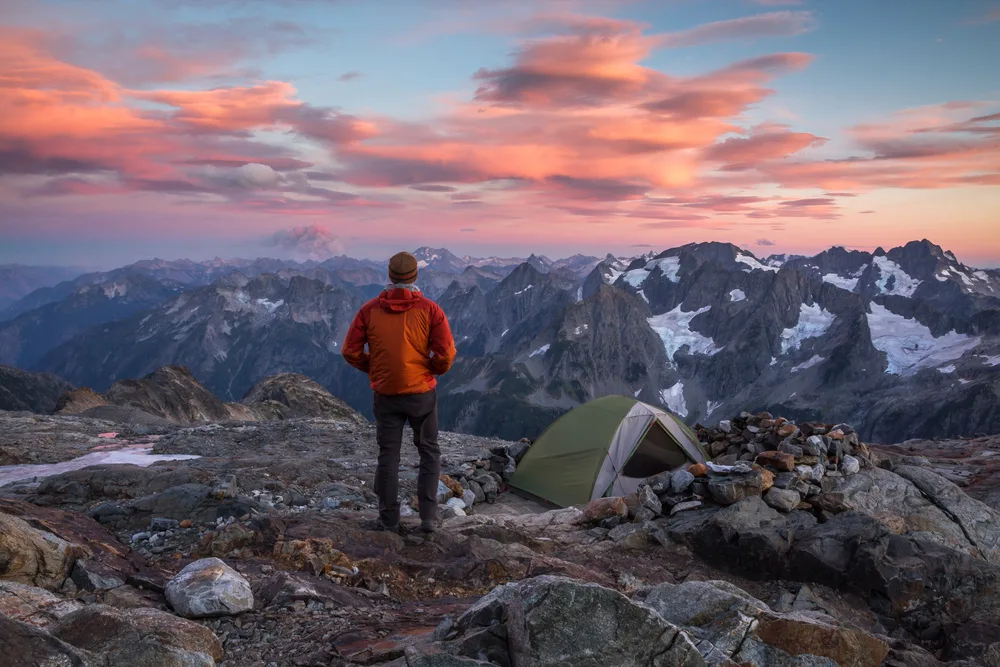 Man standing next to his tent on a mountain overlooking the valley below at dusk with an orange sky in North Cascades National Park, one of the best day trips to take from Seattle