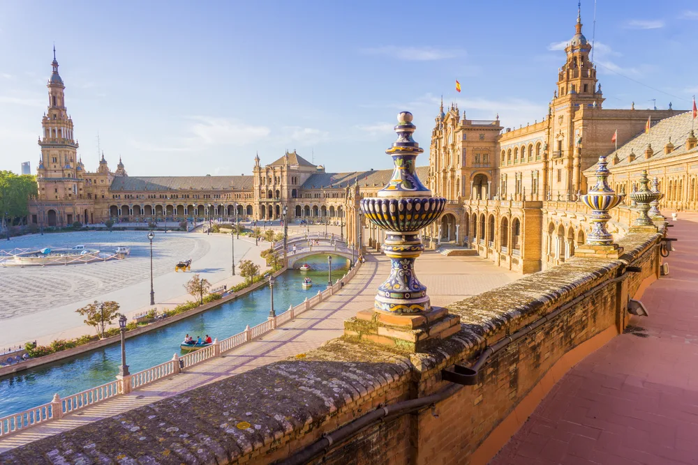 Pictured as one of the must-visit places in Spain, a photo of the Plaza de Espana in Seville with a canal running through the courtyard of a giant building complex