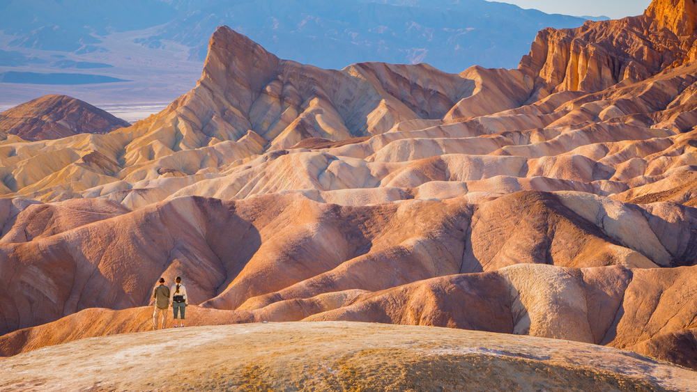 For a roundup of the best places to visit in Southern California, a pair of hikers stand in Death Valley National Park, in front of the large rock formations that look like land-locked waves