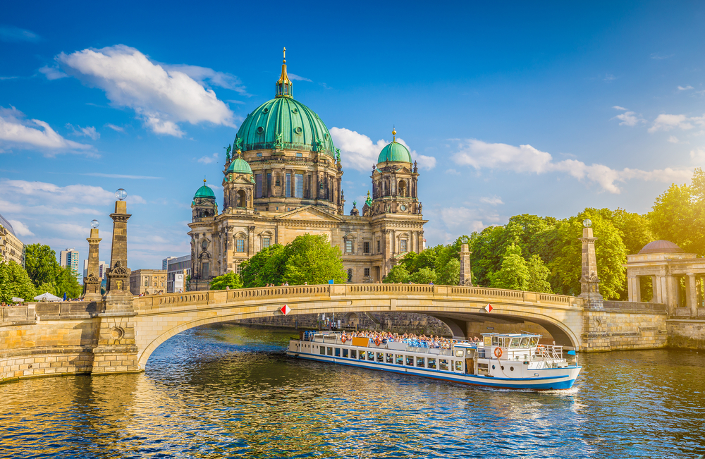 For a roundup of the best places to visit in Germany, a nice photo of sun shining on the historic cathedral and a boat traveling on the river below the stone bridge