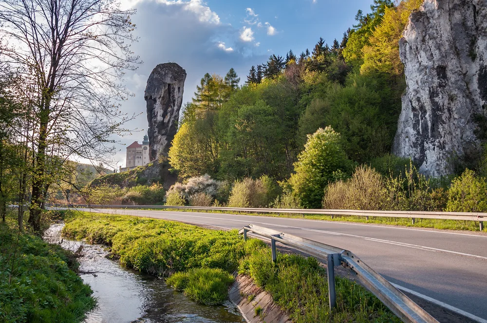 Large rock formation in Ojców National Park, a must-see place in Poland, pictured with a road running in front of it and next to a still stream
