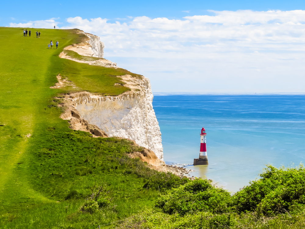 White chalk cliffs of Seven Sisters Country Park, one of the best day trips from London, as seen with green grass and a lighthouse far below