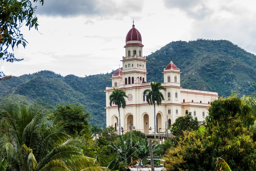 Old church in El Cobre Village, Cuba with green mountains and clouds in the background for a piece showing how Americans can legally visit Cuba