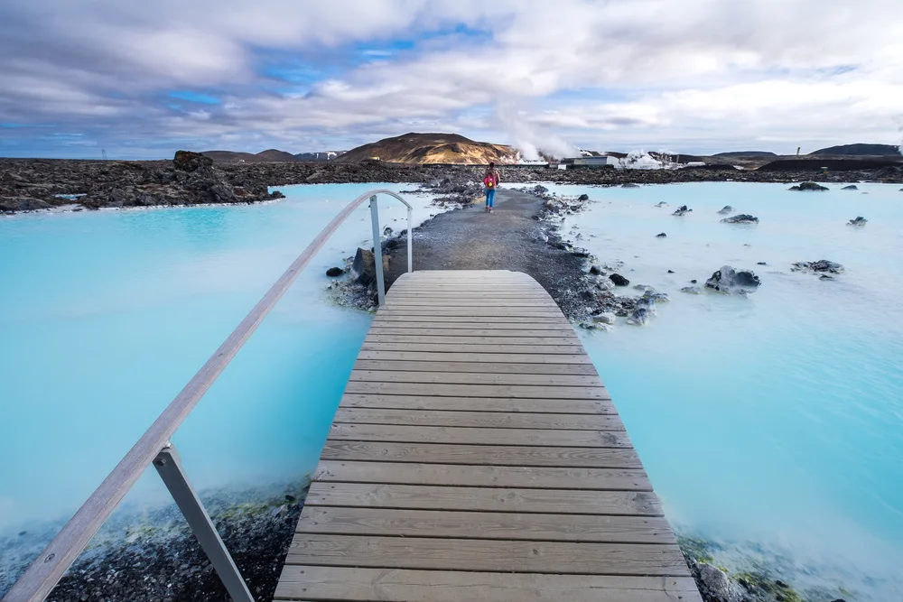 Woman in a red shirt and blue pants making her way down a rocky path, just across the wooden bridge that crosses the blue lagoon