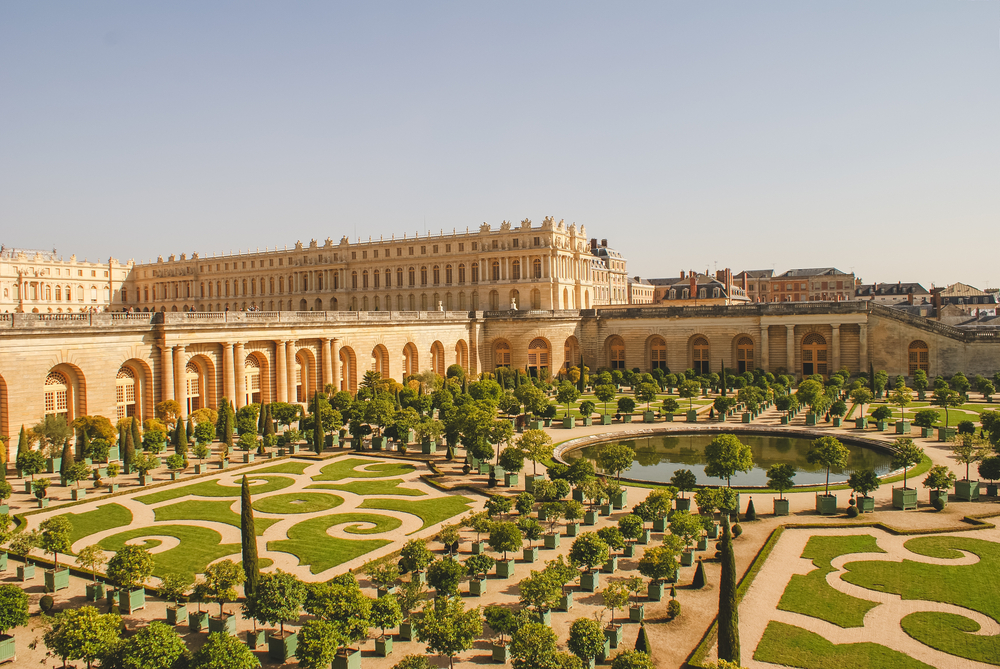 Exterior of the Royal Palace of Versailles pictured with its lush green gardens spread out all over in front of the palace itself