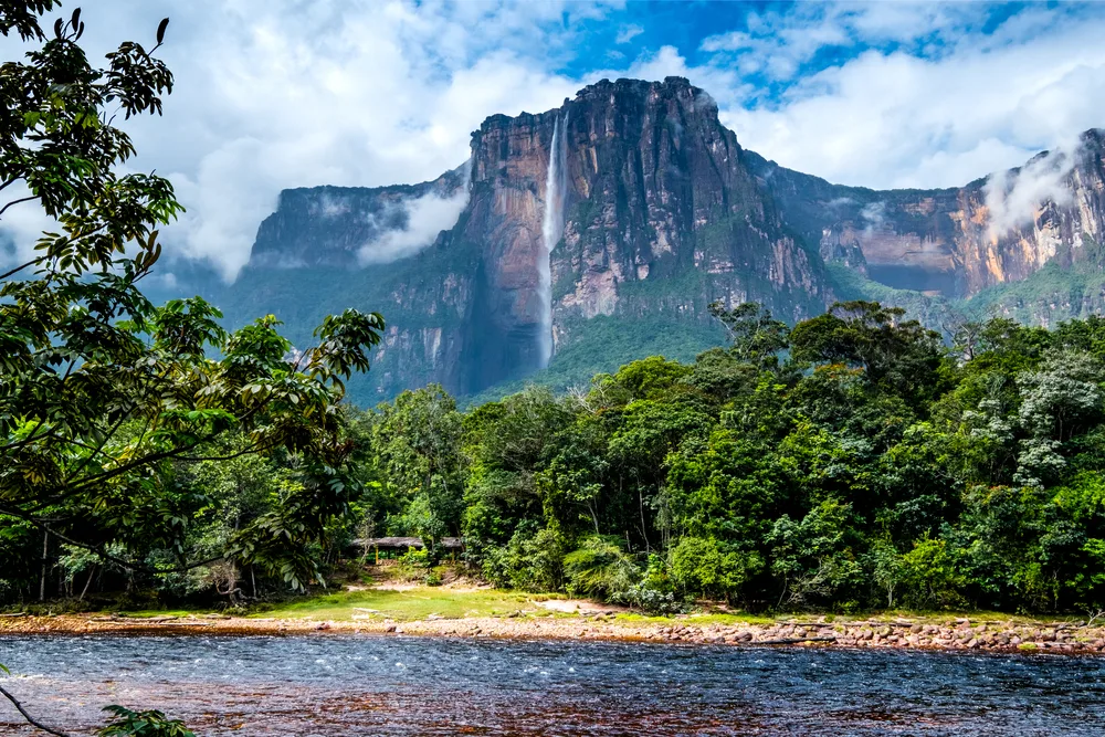 Photo taken from the banks of the river of Angel Falls, with its towering rock face and large, skinny waterfall, pictured during the best time to visit Venezuela