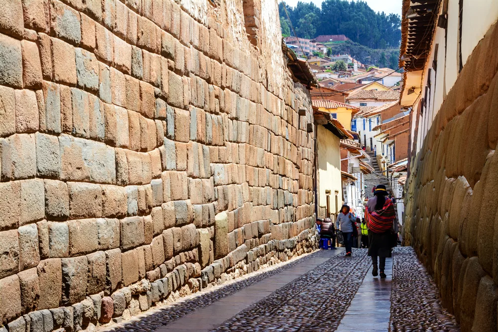 Peruvians in colorful dress walking along a narrow path between two giant stone walls in Cusco, one of our top picks for must-visit places in Peru