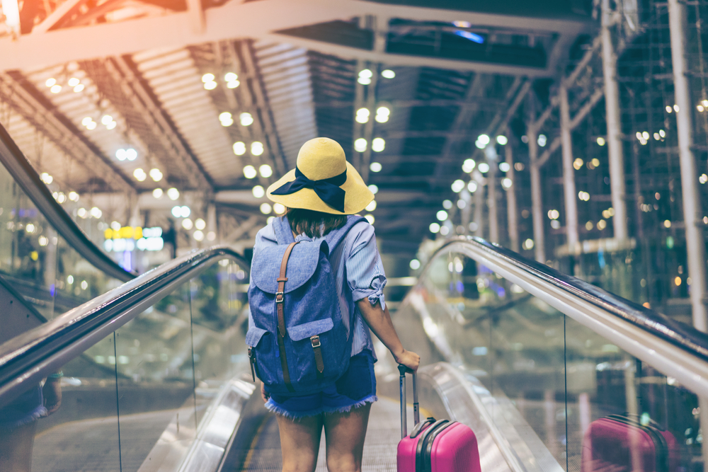 Rear view of woman in airport shows example of the best travel backpacks for women carrying a blue pack and pink luggage through the airport with a hat on