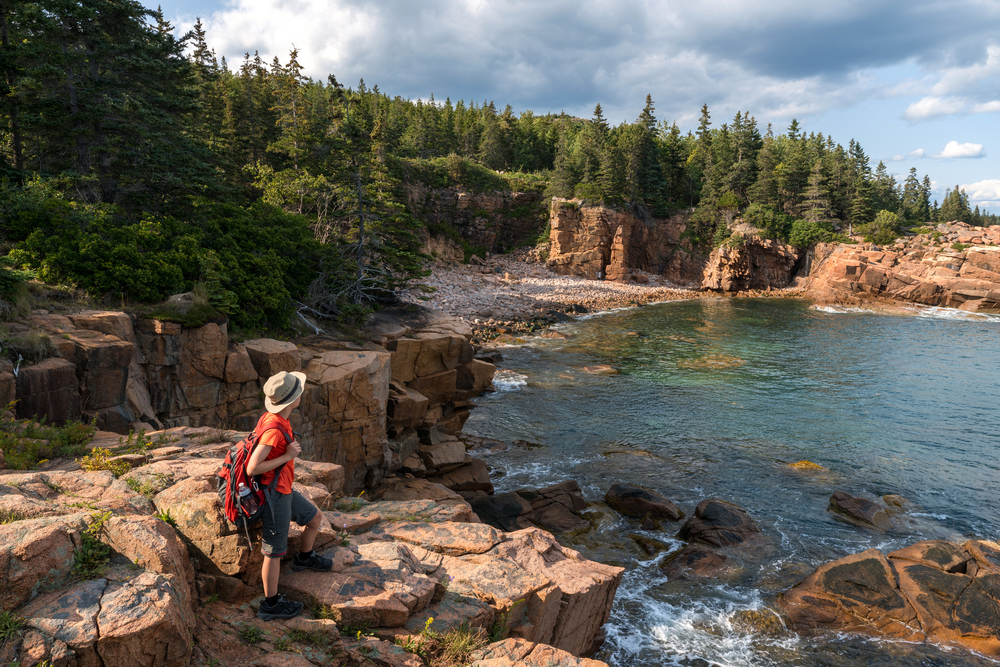 Older woman in a red shirt and shorts standing on the edge of the coast in Acadia National Park