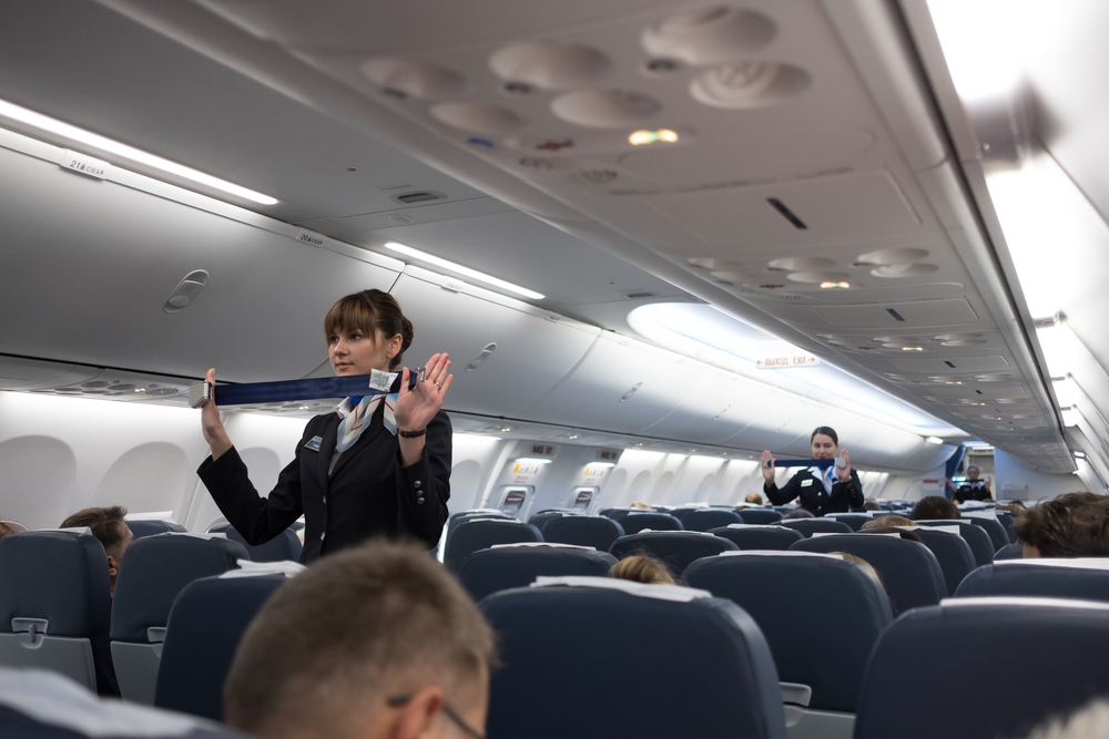 Flight attendants show travelers how to fasten seatbelts for a piece asking how safe is flying
