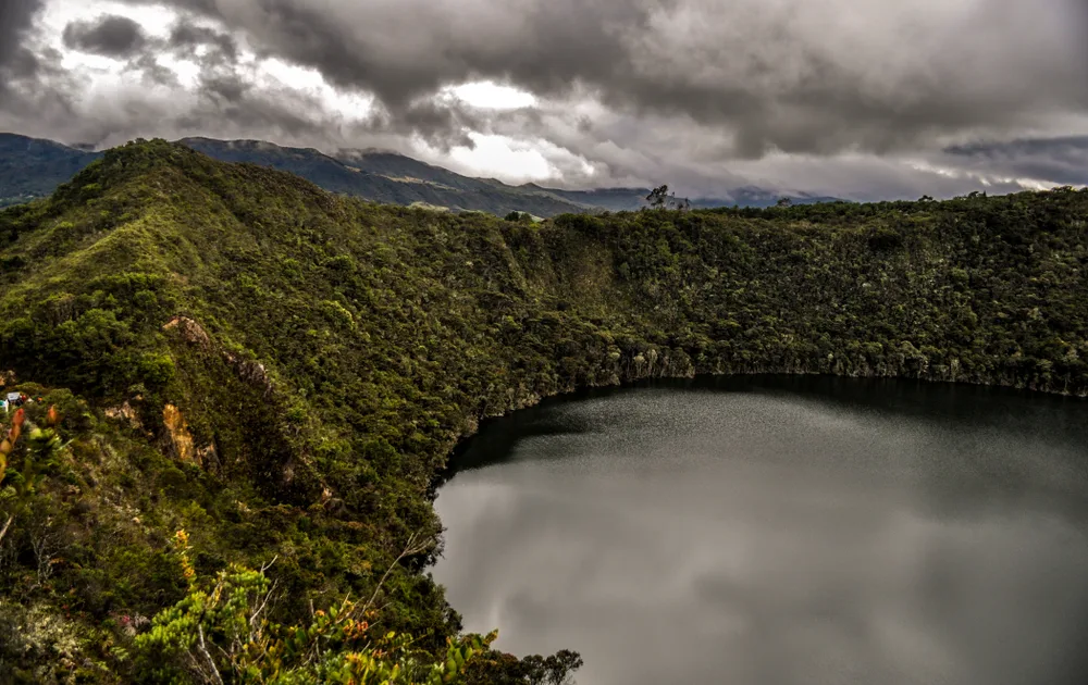Close-up of Lake Guatavita in Colombia with dark clouds overhead and still water in the crater far below the POV of the reader