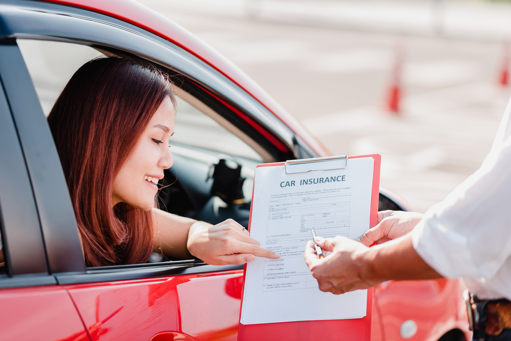 Young woman smiles and points to her car insurance policy in a red car to answer the question should you get insurance for a rental car when you have car insurance
