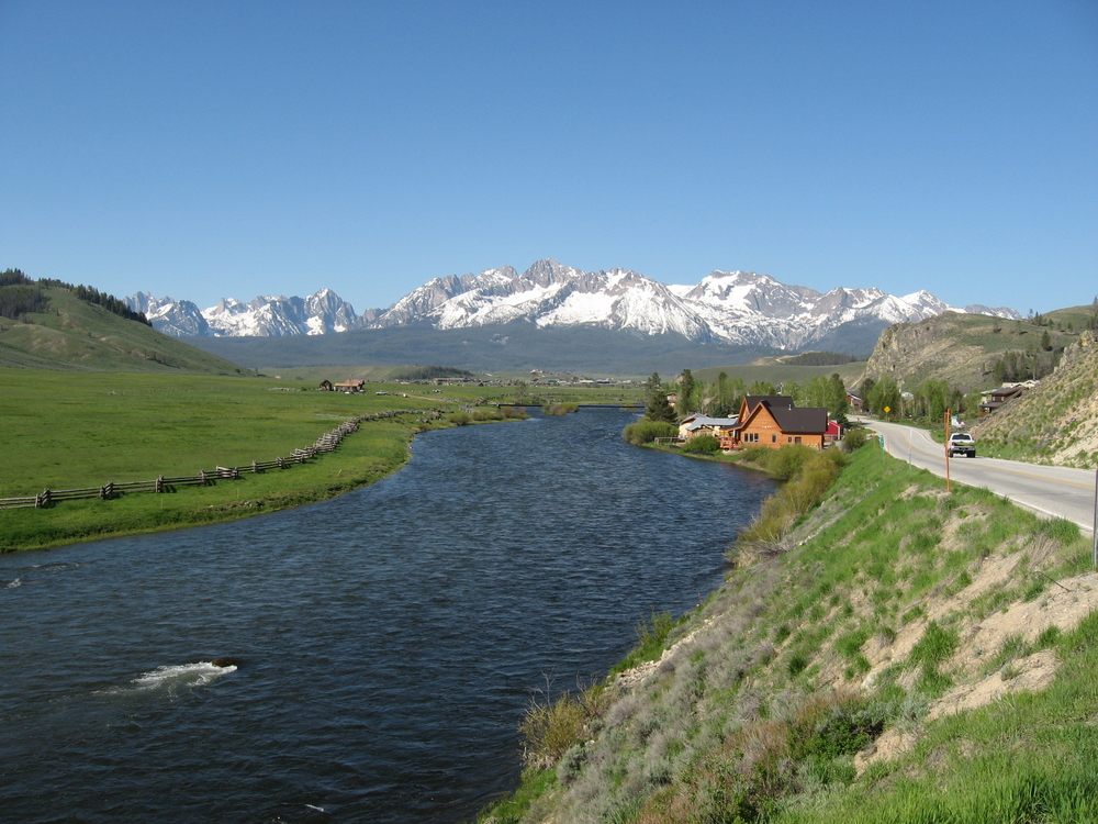 River running along a green grass pasture and a wooden fence in the foreground of a large landscape with large mountains in the background and a truck driving by in Stanley, Idaho