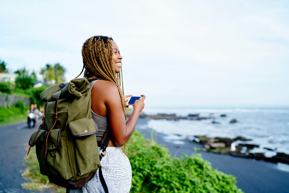 Female traveler smiles holding her smartphone while hiking with a backpack and using one of the best trip planning apps to navigate