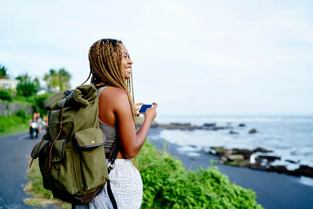 Female traveler smiles holding her smartphone while hiking with a backpack and using one of the best trip planning apps to navigate