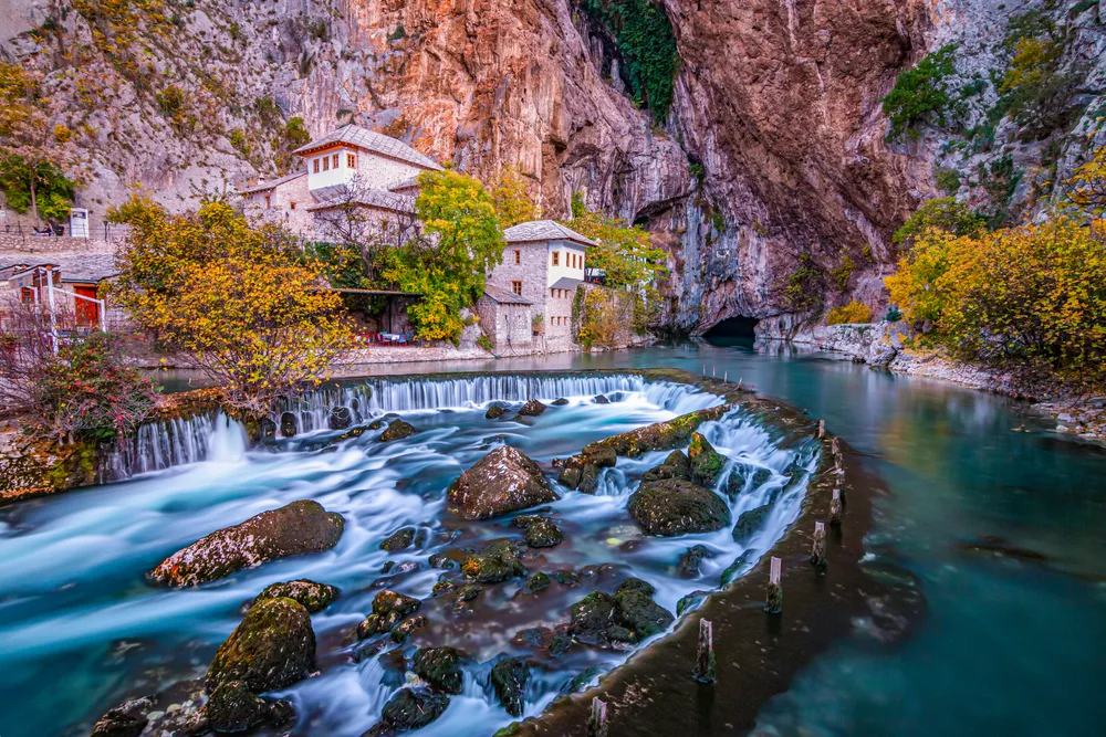 Blagaj village with rushing water over rocks with buildings in front of cliffs during fall, the cheapest time to visit Bosnia