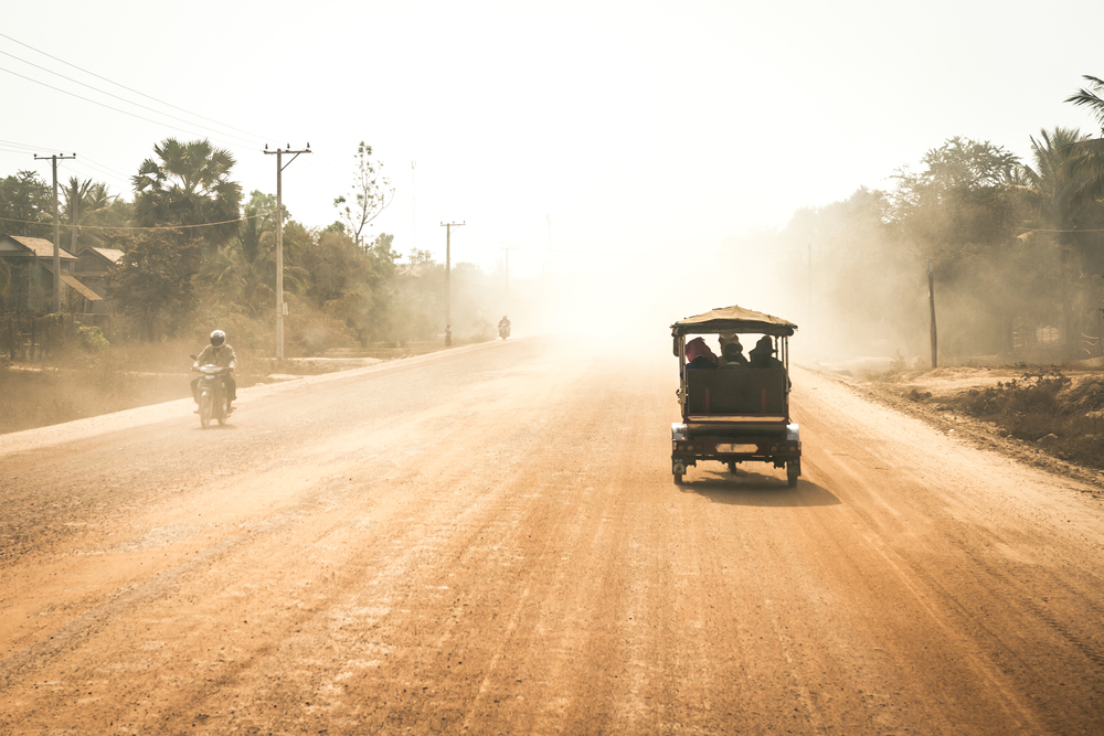 Tuktuk driving down a wide dirt road with dust flying all over the town and a motorcycle going the opposite way during the off-season, the cheapest time to visit Cambodia