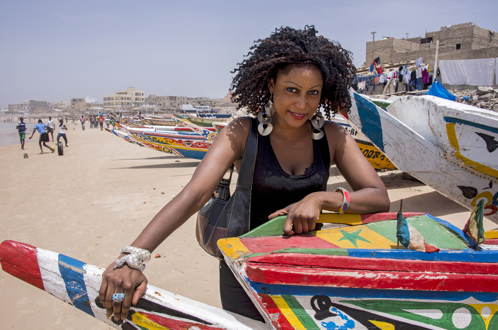 Woman smiles and stands next to a colorful fishing boat in Dakar for a piece titled Is Senegal Safe to Visit