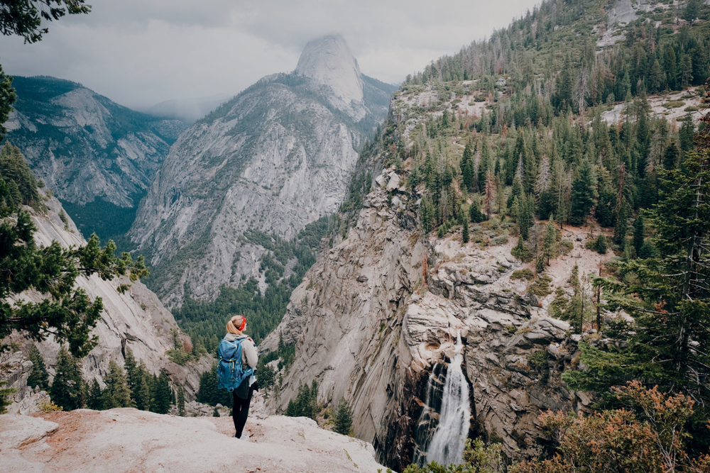 For a piece titled is Yosemite safe to visit, a woman with a blue backpack hiking on the side of a mountain overlooking the snowy and frozen-waterfall valley below