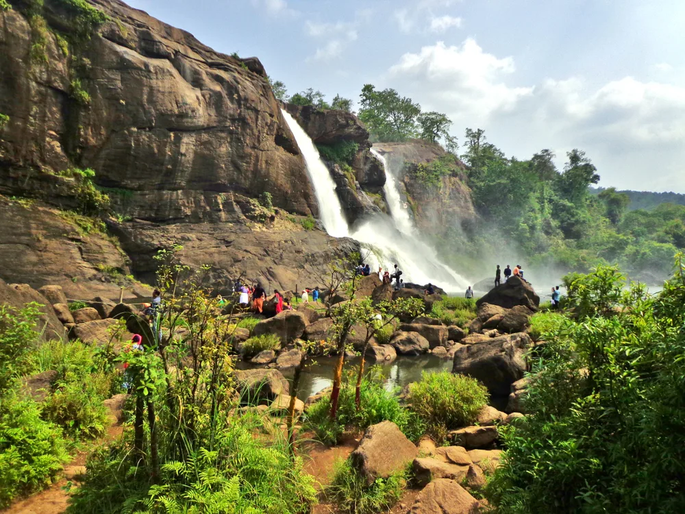 Gorgeous Athirapally Waterfalls in Kerala's jungle pictured on a clear day with vegetation between it and the camera pictured for a piece titled Is Kerala Safe to Visit