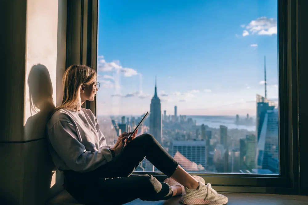 Blonde woman sits in the windowsill of a high-rise apartment in New York City, using a tablet to see one of the best travel websites for ideas on where to travel next