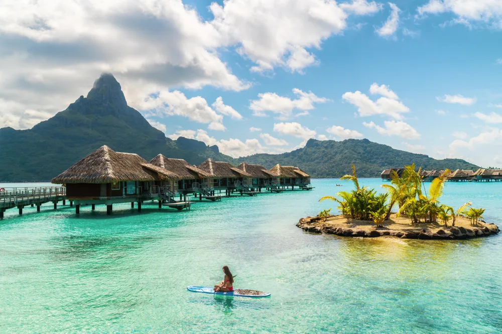 Pretty and thin Asian woman on a paddleboard floating between huts and an island for a piece titled Is Bora Bora Safe to Visit this year