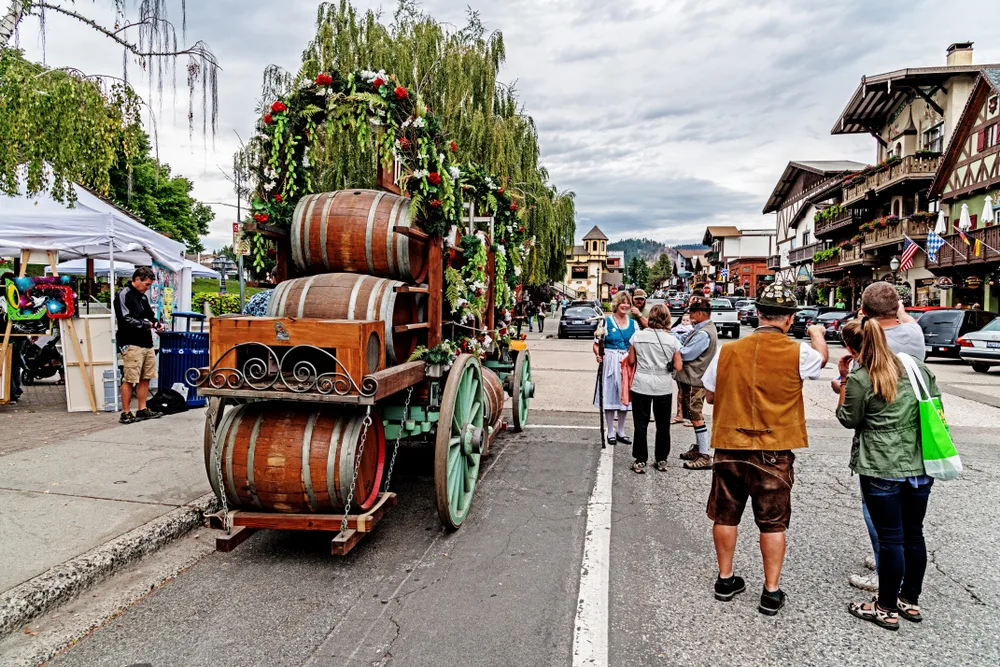 People in classic Bavarian dress walking by an old-time wagon with wooden barrels on it in the heart of downtown, the best area to stay when visiting Leavenworth WA