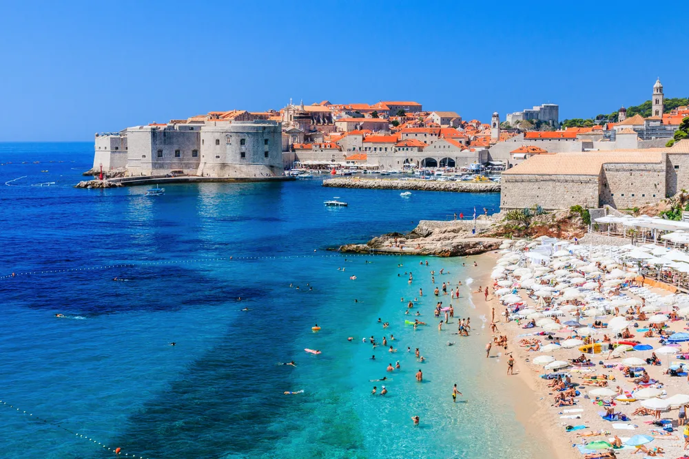 View of the sandy beach with turquoise water and tourists on the shore in front of the fortress walls of the Old City during the best time to visit Dubrovnik