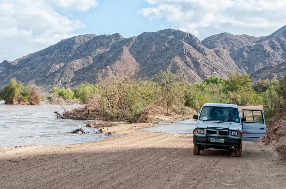 A blue 4x4 driving by the pond oasis in Namibia for a piece on whether it's safe to visit the area with the Sendelingsdrift mountains in the background