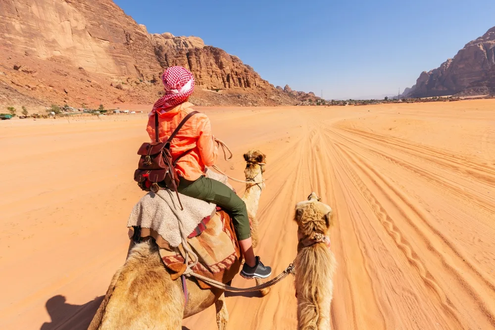 Beautiful woman riding a camel in a headscarf pictured in the middle of a desert with tan sand