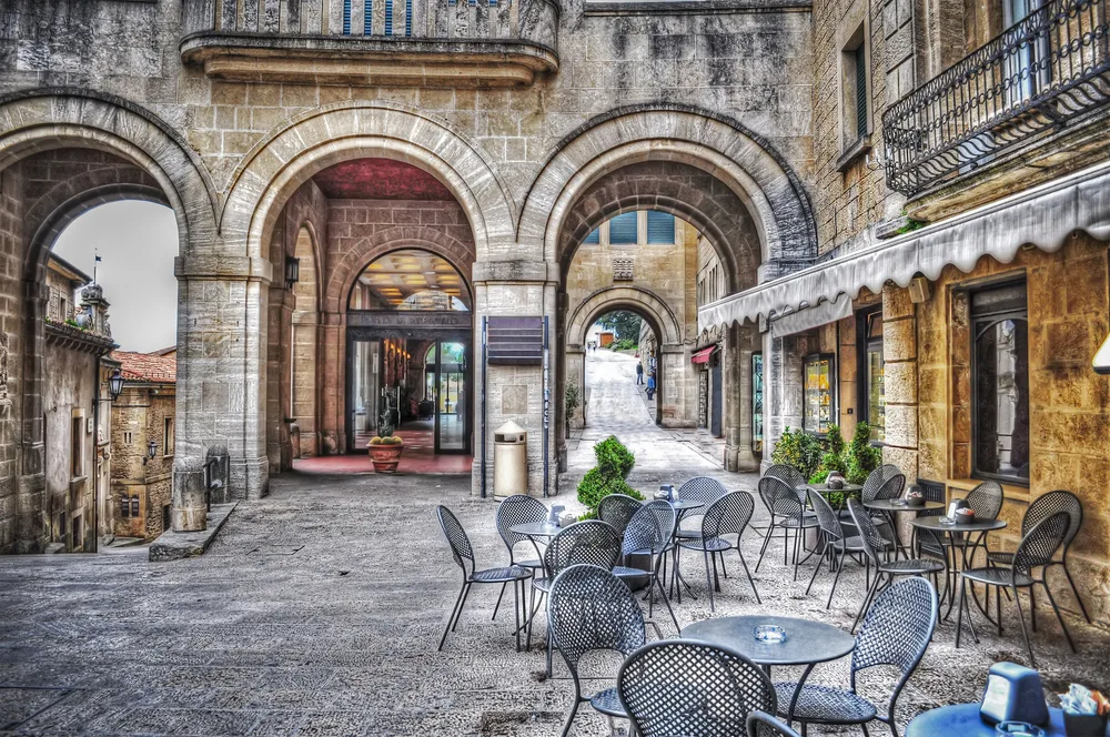 Outdoor dining table and chairs in front of Romanesque arches in a public square to show why you should visit San Marino