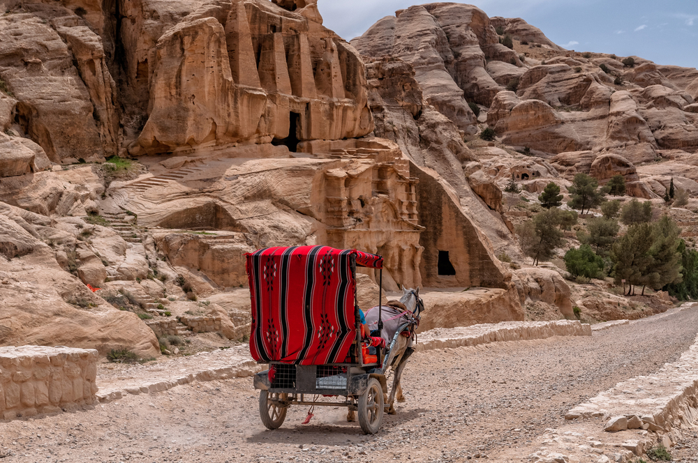 A horse-drawn carriage with a red blanket draped over it making its way down the gravel path near the entrance to the famous Petra cliffside ruins