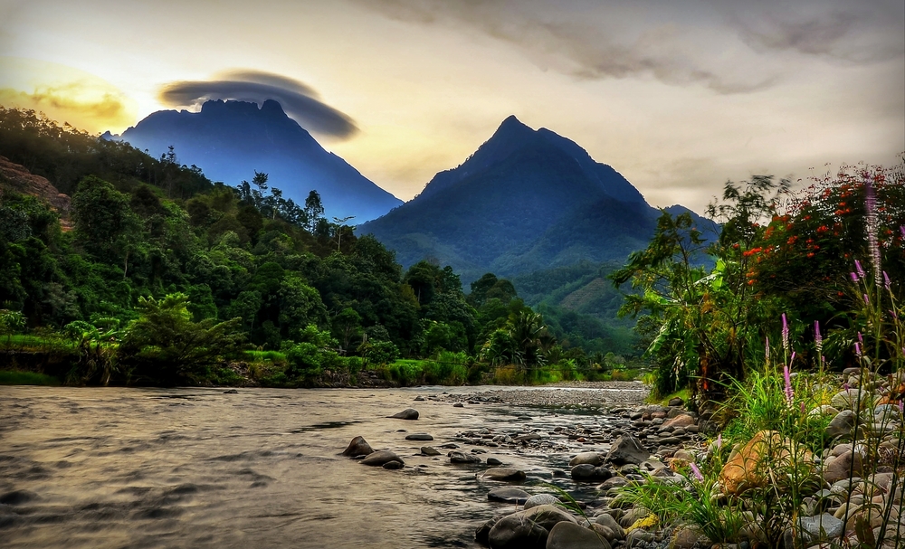 Village of Tambatuon in Borneo for a piece titled Is Borneo Safe to Visit with a cloud-capped mountain towering over a stream with water running down the rocky area