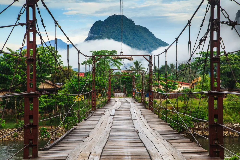 Gorgeous natural beauty seen from a wooden bridge over a jungle with a mountain in the background for a piece titled Is Laos Safe to Visit