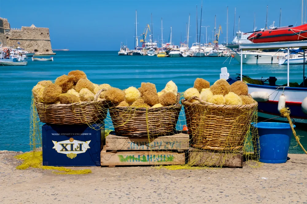 Photo of several sponges sitting in big wicker baskets next to the ocean for a piece on when to visit Greece to get the best deal and avoid crowds