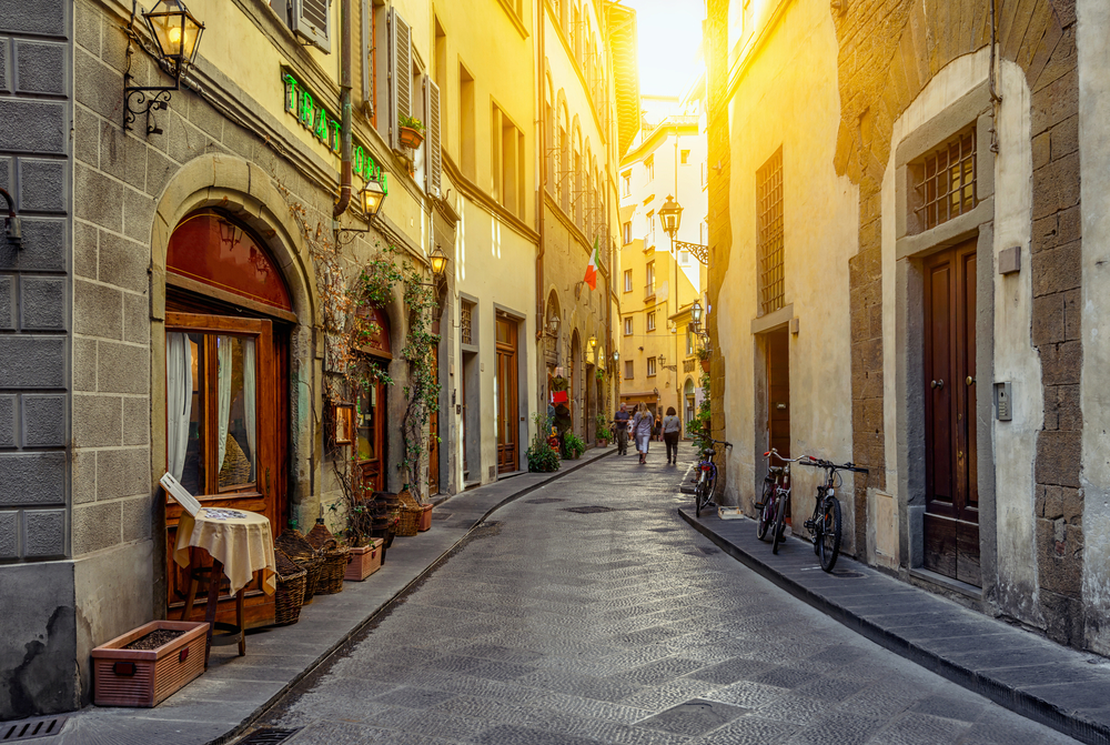 A narrow street winding through the city with beautiful architecture on either side and the setting sun shining through during the least busy time to visit Florence