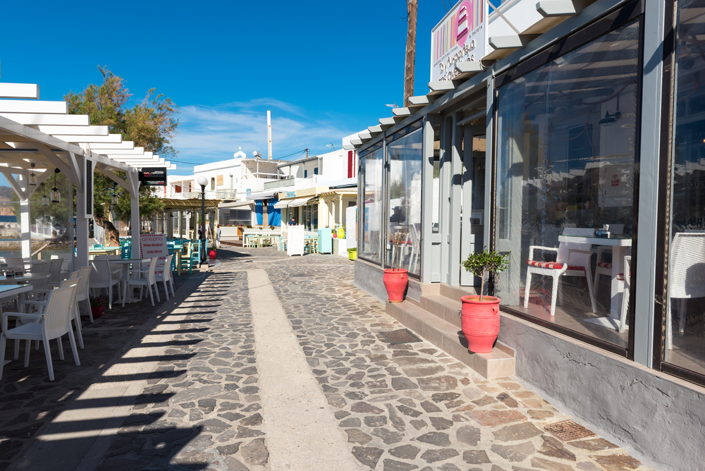 Small white restaurants and shops line the cobblestone street in Pollonia, one of our favorite places to stay in Milos, pictured under a light blue sky