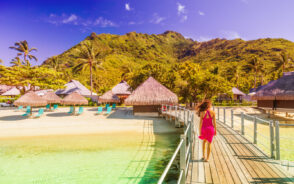 Woman in a pink dress walking along the elevated bridge boardwalk toward the luxury huts below an orange sky and above teal water for a piece titled Is Tahiti Safe to Visit
