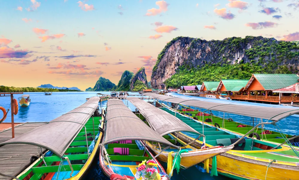 Bright colored boats floating on the water on the beach while the golden sunset falls over the ocean for a piece titled Is Phuket Safe to Visit