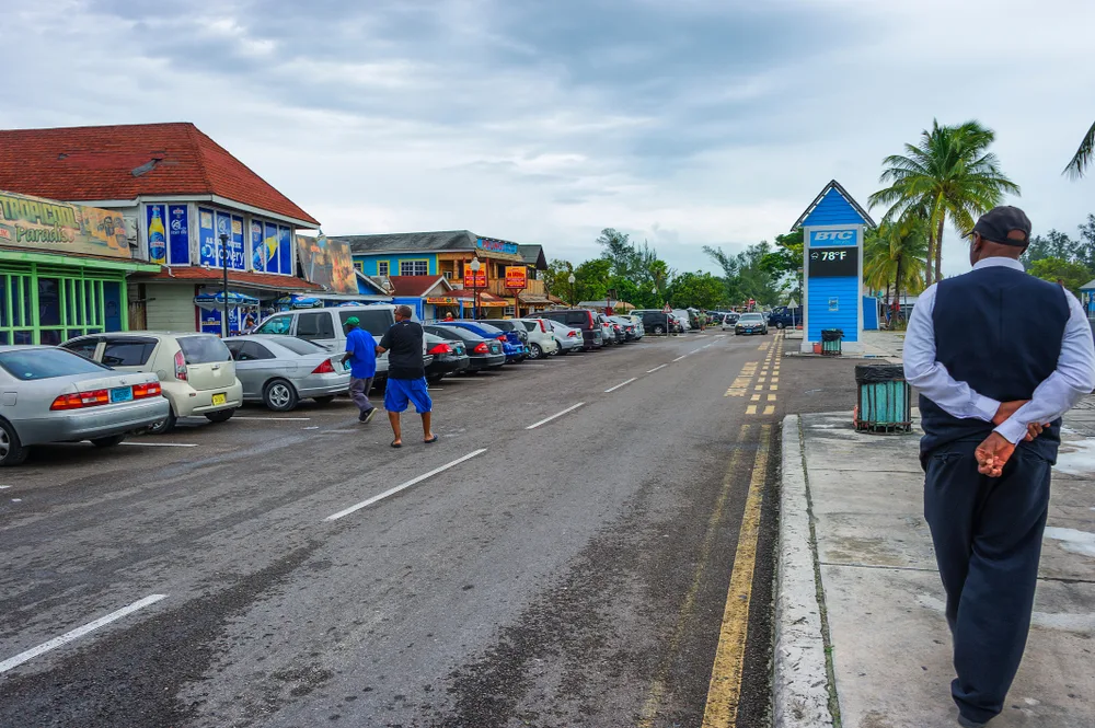 Man walking with his arms behind his back looking across the street at the colorful buildings that house the Fish Fry, one of the most unsafe areas in Nassau