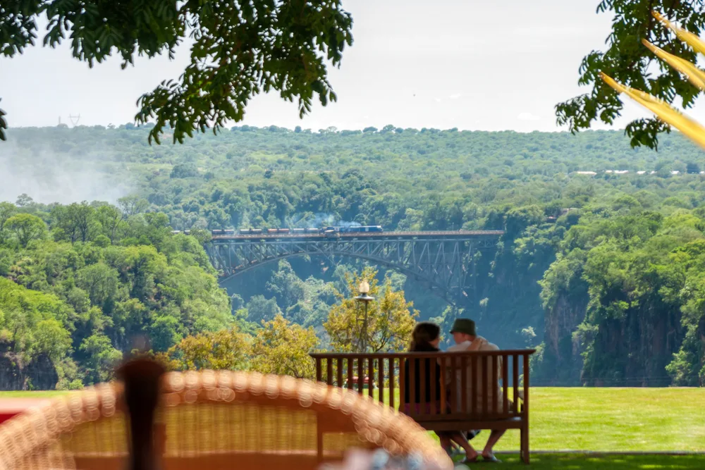 Image of a couple sitting on a park bench overlooking a bridge with Victoria Falls in the background for a piece on whether or not the falls are safe to visit